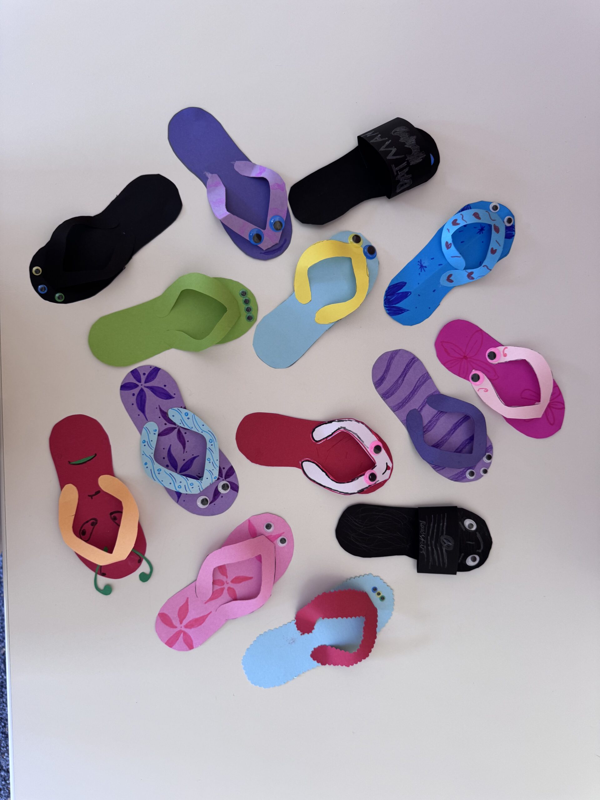 Bunte gebastelte Papier-Flip-Flops mit aufgeklebten Kulleraugen und gemusterten Fußriemen auf weißem Untergrund