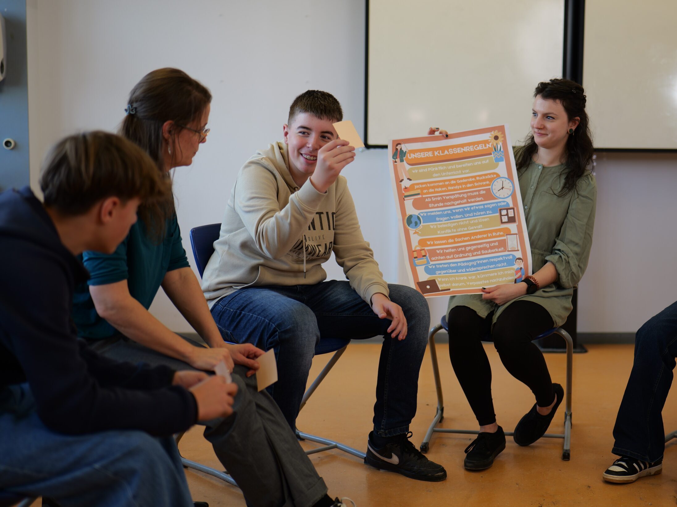 Eine Gruppe Jugendlicher sitzt im Stuhlkreis; eine junge Frau hält ein Plakat mit der Überschrift Unsere Klassenregeln hoch