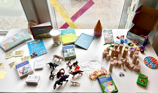 viele verschiedene Materialien wie Stifte, Holzfiguren, Tierfiguren und Schreibutensilien liegen bunt verteilt auf einem Tisch vor einem Fenster.