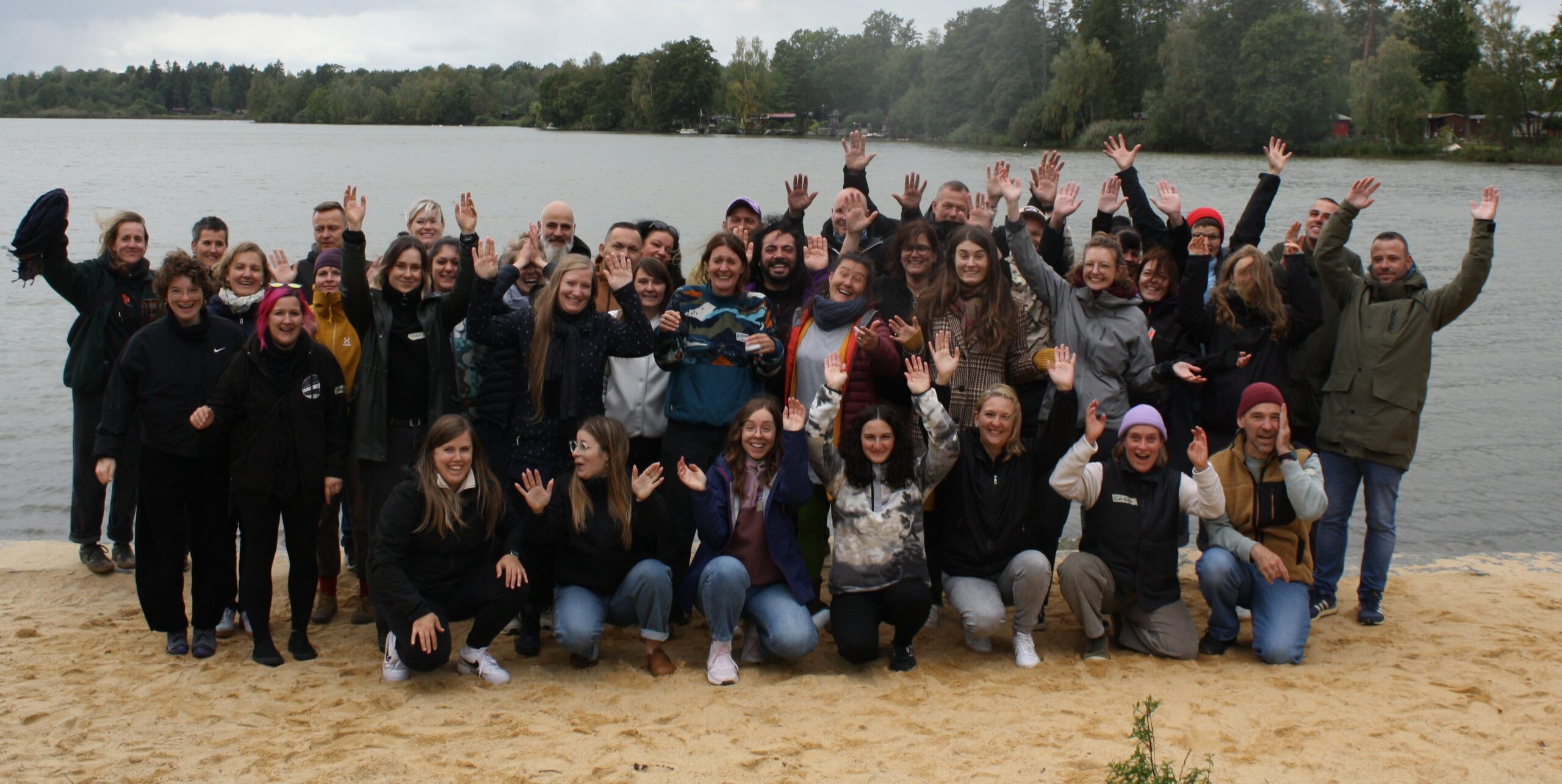 Große Gruppe von Personen steht und kniet am Ufer eines Sees für ein gemeinsames Gruppenfoto. Viele Personen haben die Arme gehoben, im Hintergrund sind Wasser, Bäume und ein bewölkter Himmel zu sehen